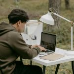 Seorang pria duduk di kursi tengah hutan sambil belajar menggunakan laptop dan buku, di atas meja lengkap dengan lampu belajar.